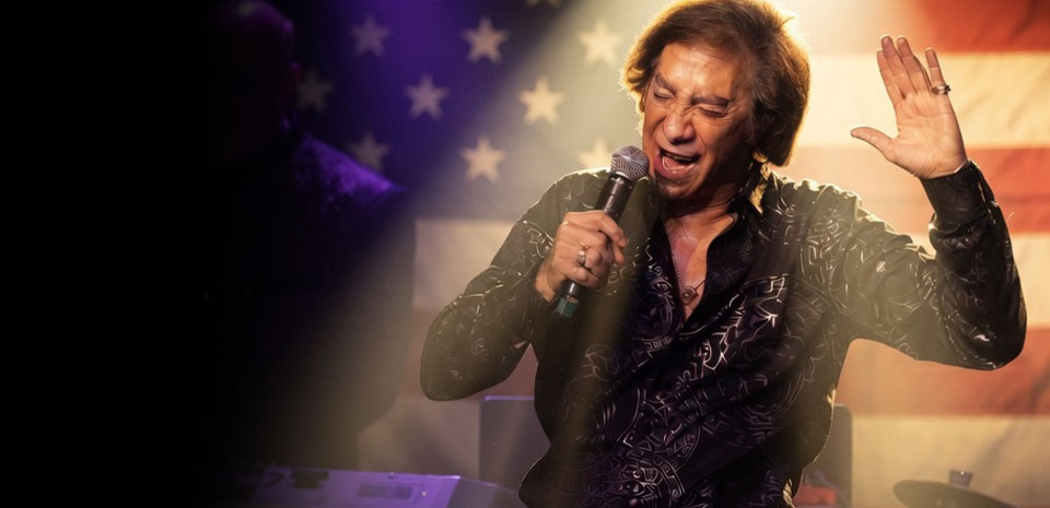 Rob Garrett as Neil Diamond singing in front of an American flag in King of Diamonds, Las Vegas